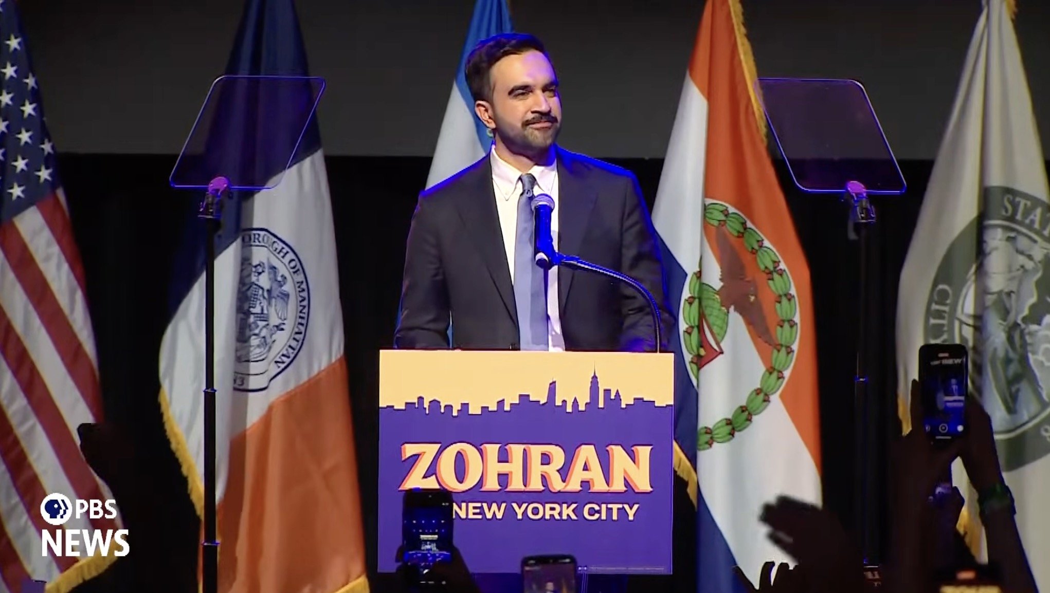Speaker at a podium during a political event in New York City, with flags in the background and audience members raising their hands.