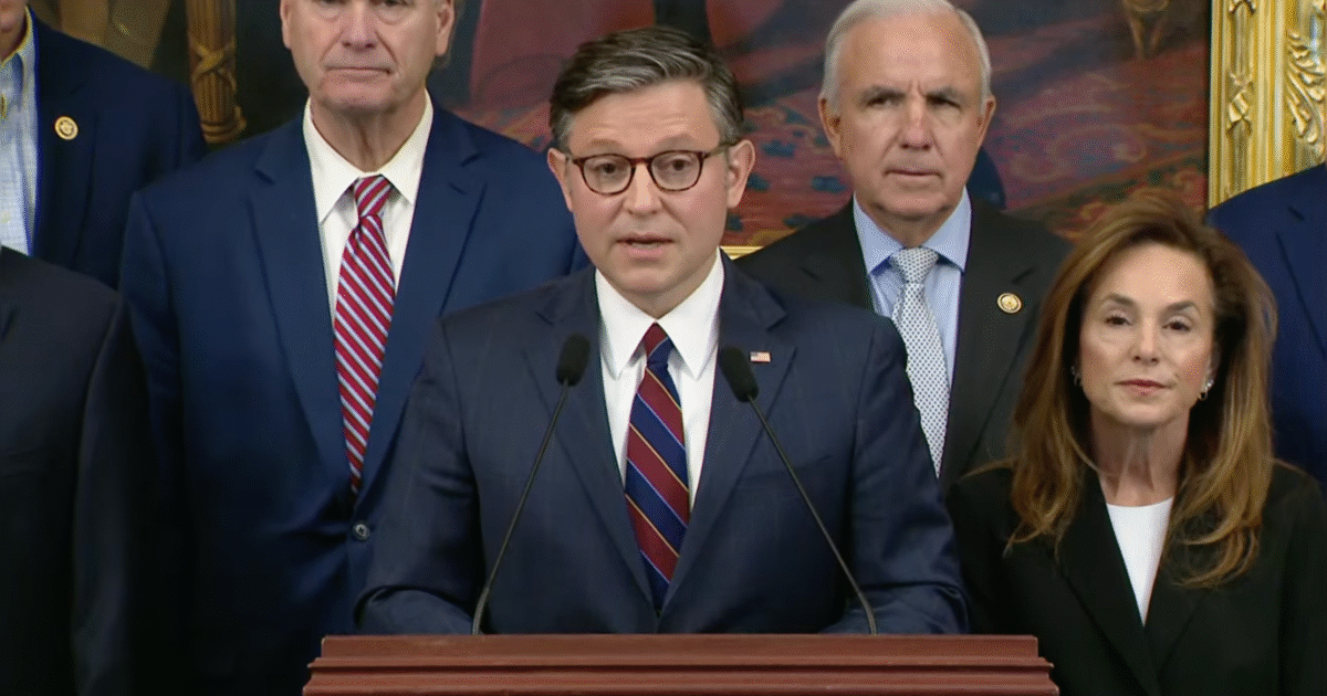 A group of politicians stands behind a speaker at a press conference, discussing important legislative issues in a formal setting.