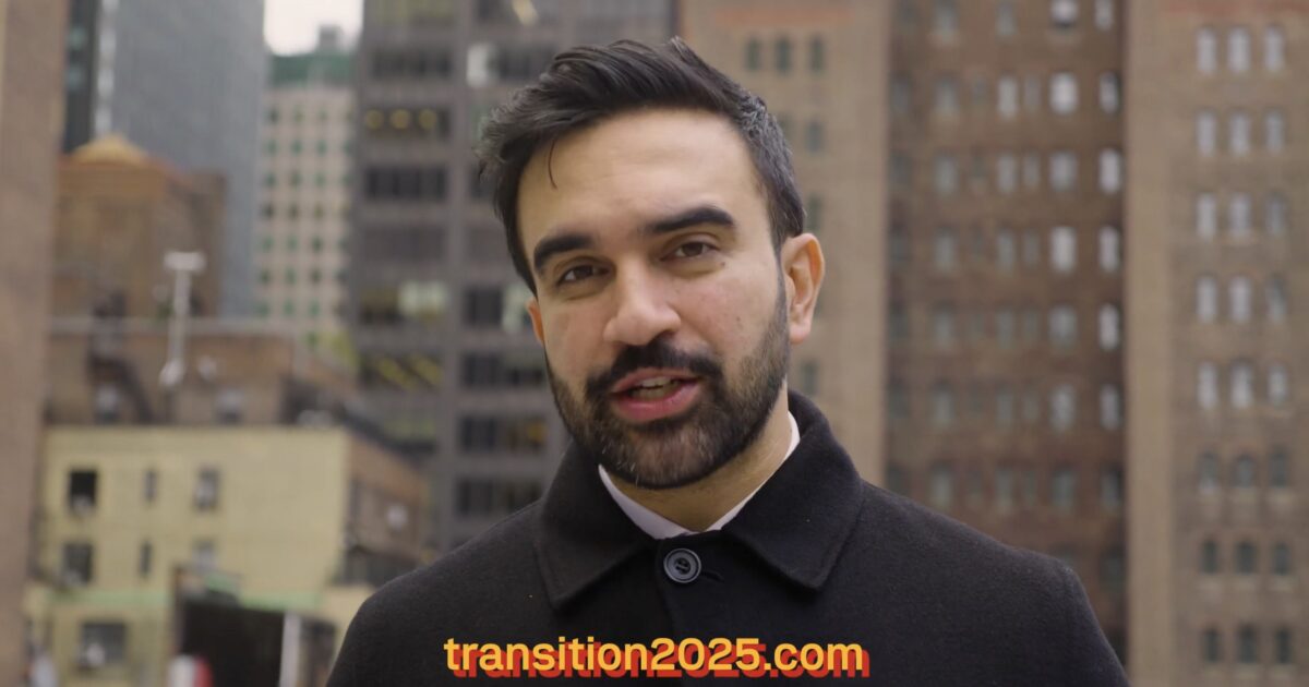 Man in a black coat speaking outdoors in an urban setting, with buildings in the background and the text transition2025.com prominently displayed.