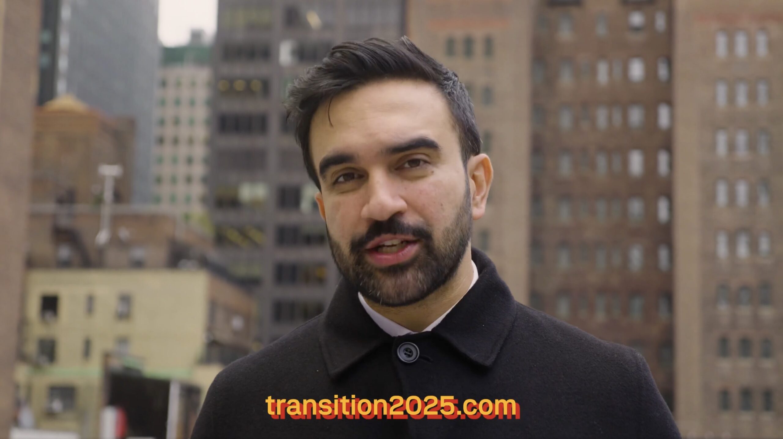 Man in a black coat speaking outdoors in an urban setting, with buildings in the background and the text transition2025.com prominently displayed.