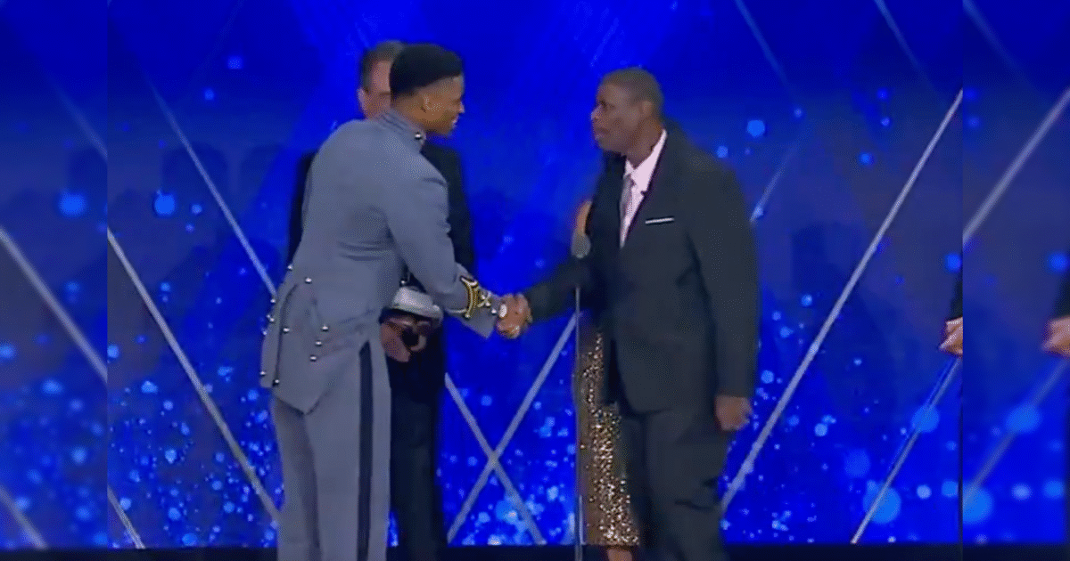 Two men in formal attire shake hands on stage during an awards ceremony, with a woman and another individual visible in the background.