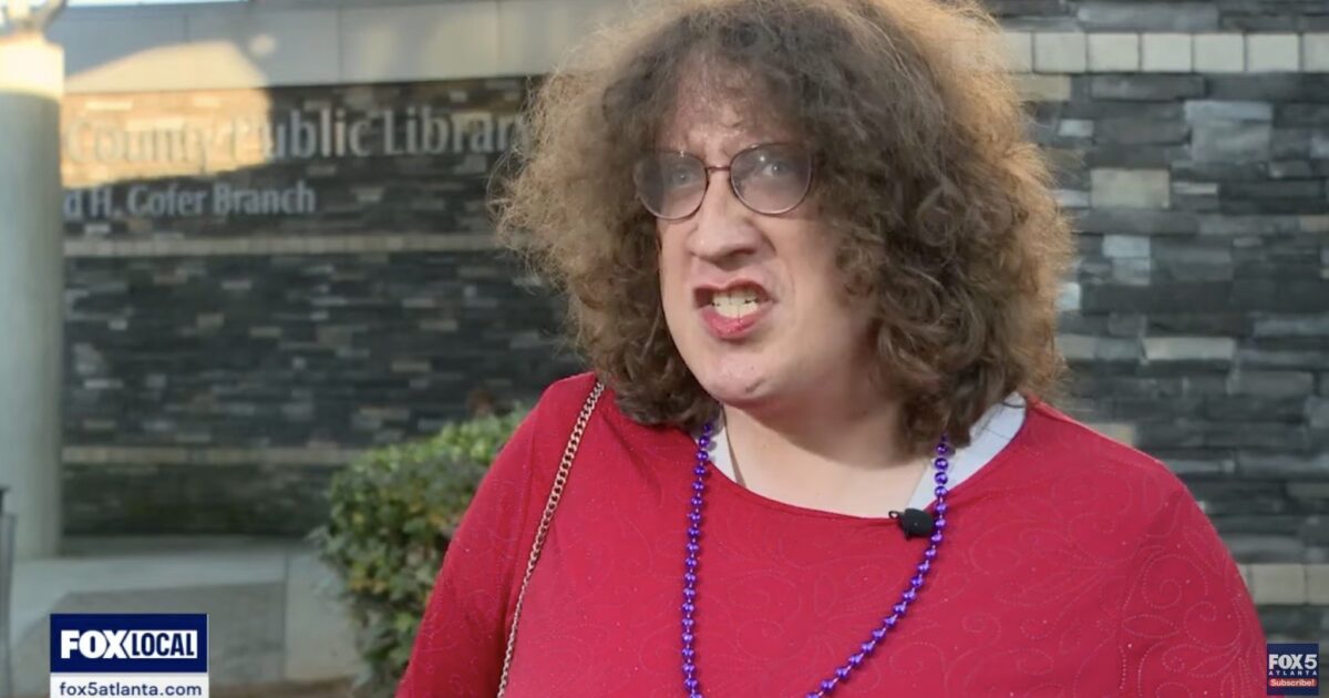 Person speaking outside a public library, wearing a red shirt and glasses, expressing strong emotions during an interview.