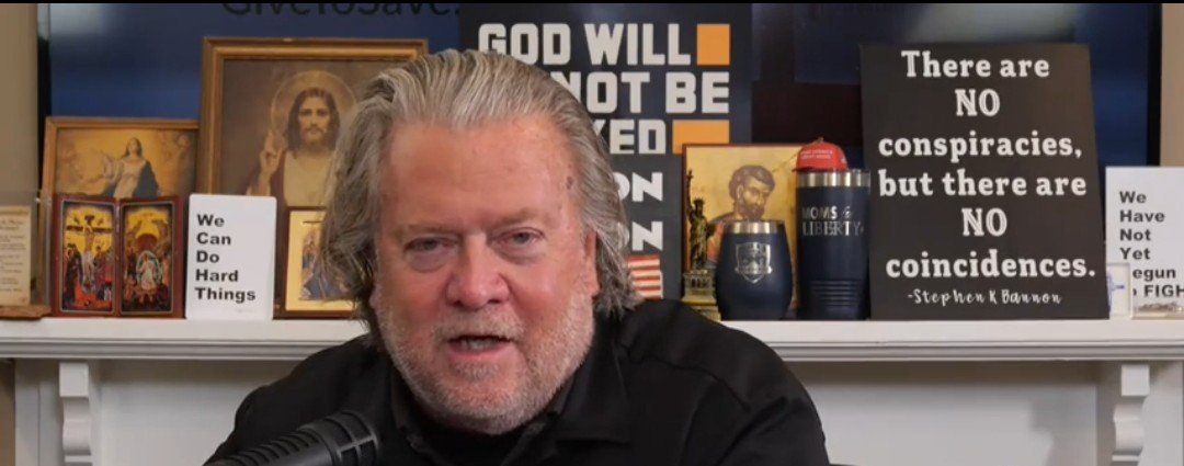 Steve Bannon speaking in front of a shelf decorated with religious icons and signs conveying motivational and controversial messages.