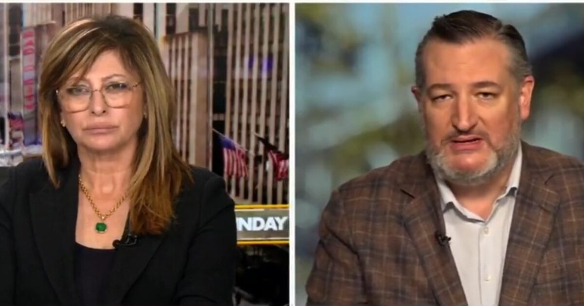 Television interview featuring a female host and a male guest discussing current events, with a cityscape background visible through a window.