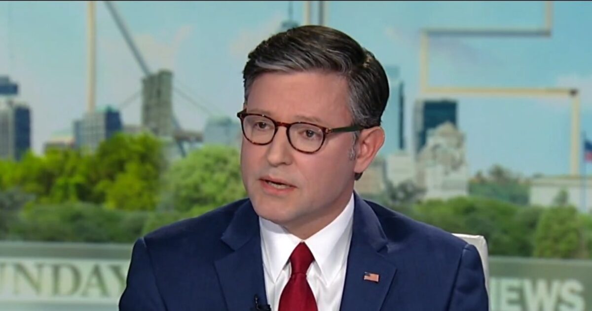 Male news anchor discussing current events on a television program with a city skyline in the background.