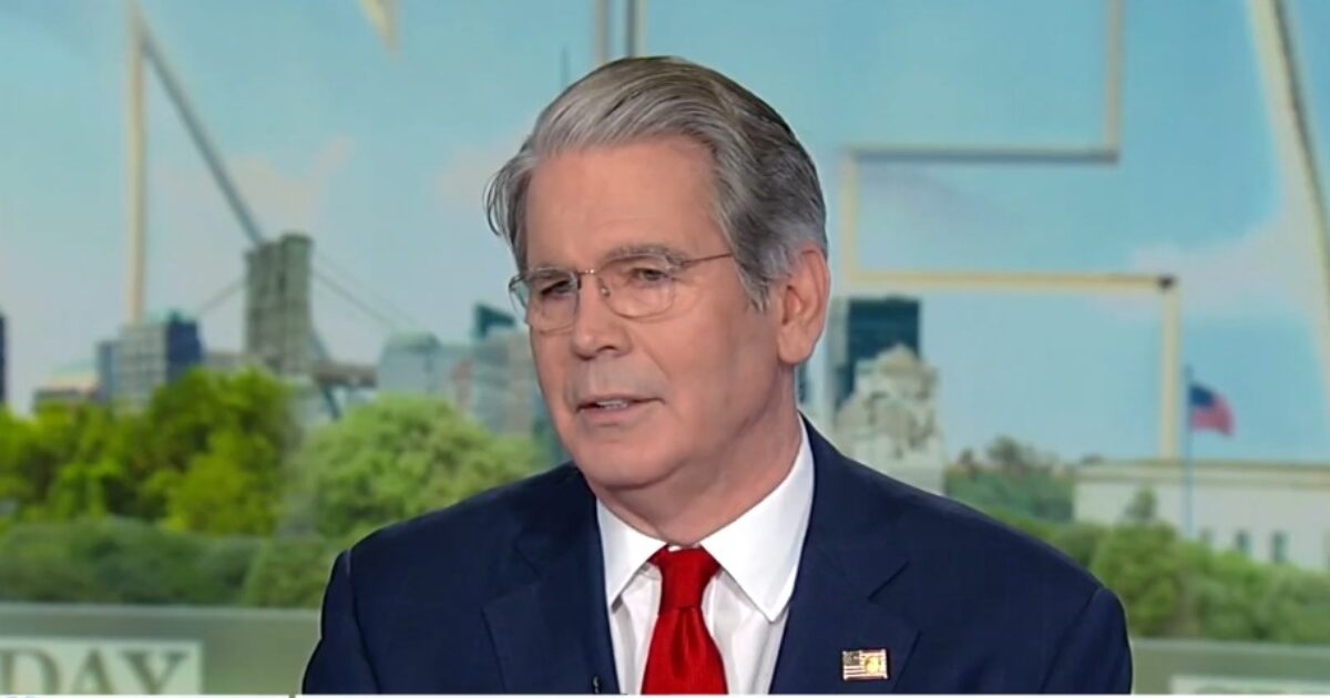 Man in a suit with glasses and a red tie speaking during a televised interview against a city backdrop.