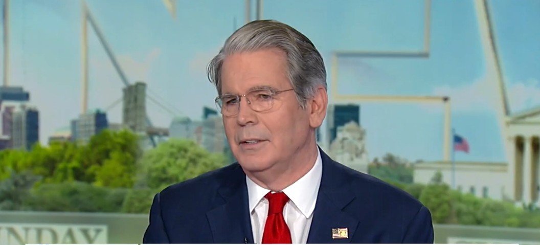 Man in a suit with glasses and a red tie speaking during a televised interview against a city backdrop.