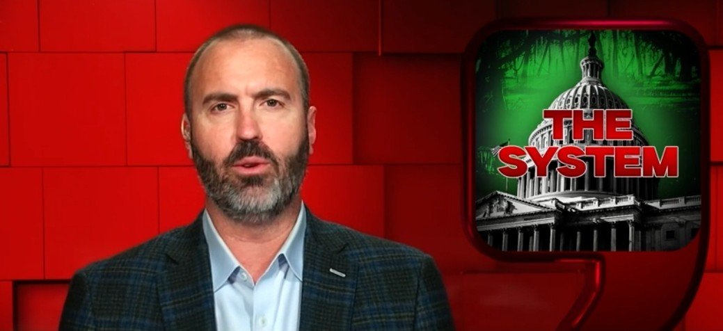 Presenter discussing political themes with a backdrop featuring the U.S. Capitol and the text "The System," highlighting issues related to governance and societal structures.