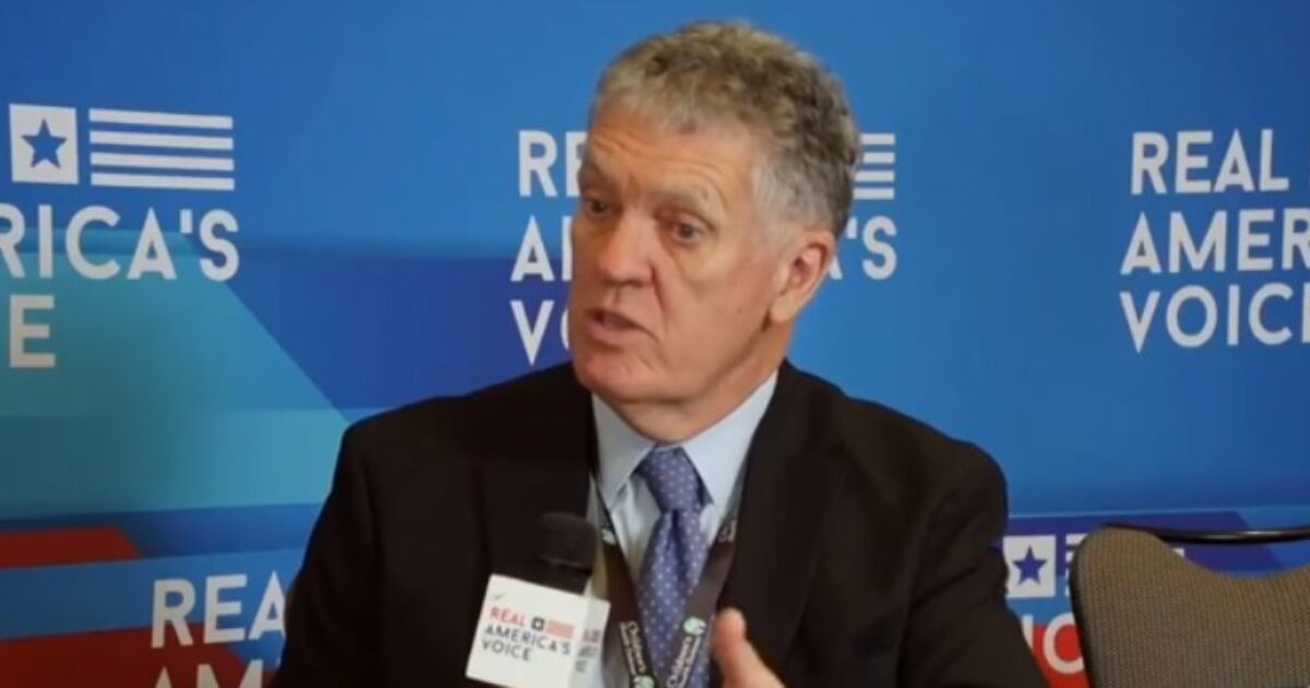 Man in a suit speaking into a microphone during an interview, with a blue and red backdrop featuring the text Real America's Voice.