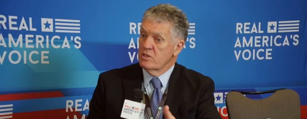 Man in a suit speaking into a microphone during an interview, with a blue and red backdrop featuring the text Real America's Voice.