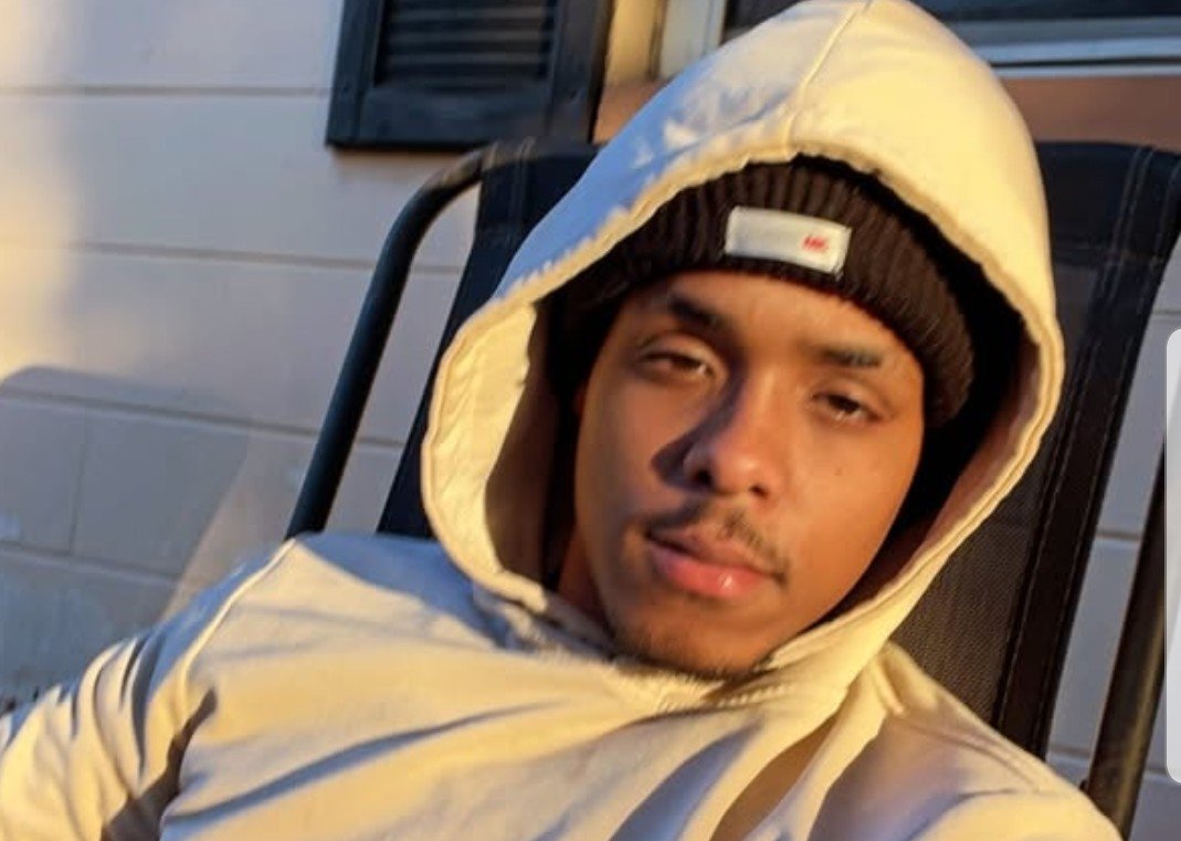 Young man wearing a beige hoodie and black beanie, sitting casually in a chair with sunlight illuminating his face.