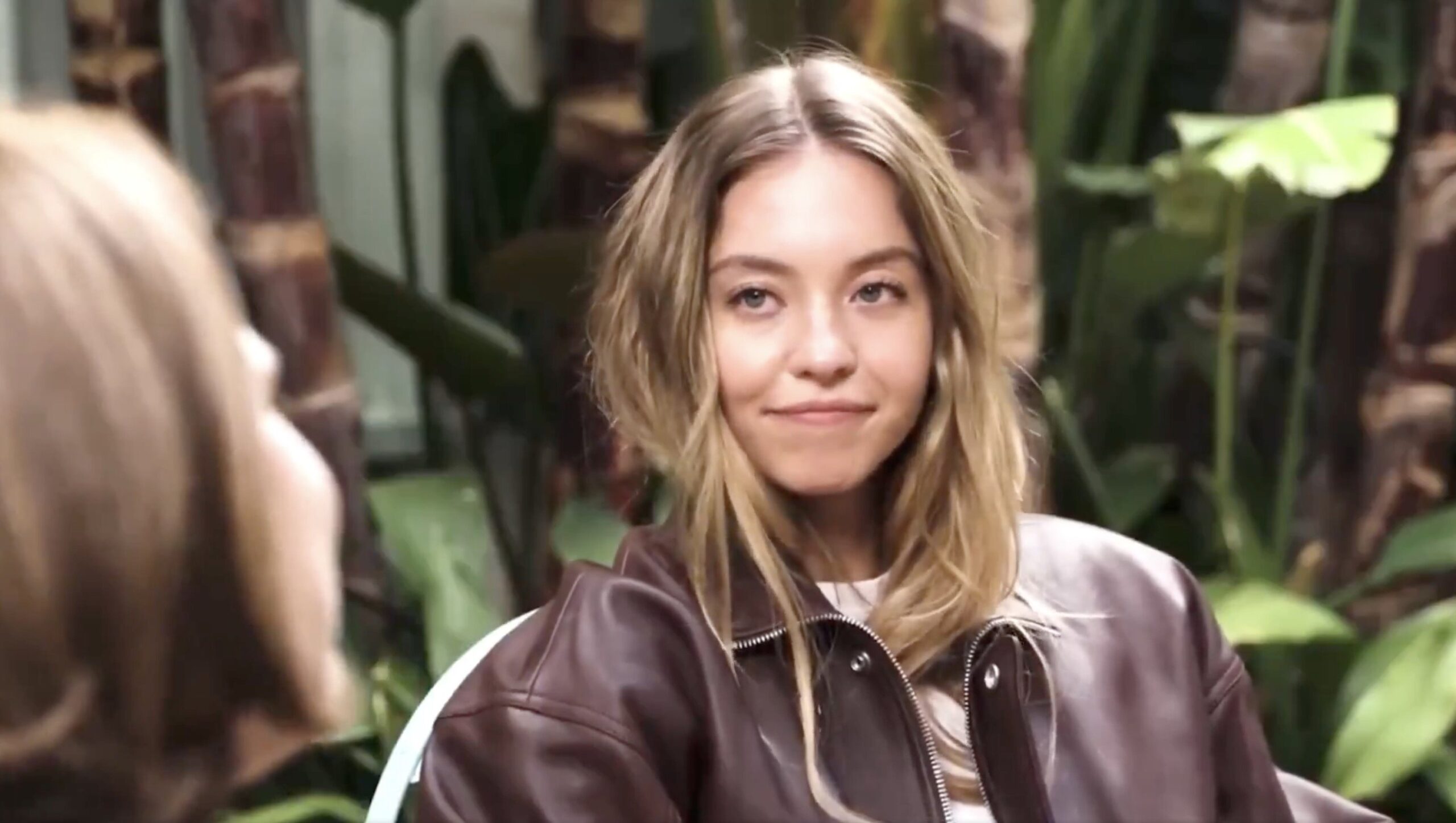 Young woman with long, wavy hair wearing a brown leather jacket, seated in a lush green outdoor setting, smiling while engaging in conversation.