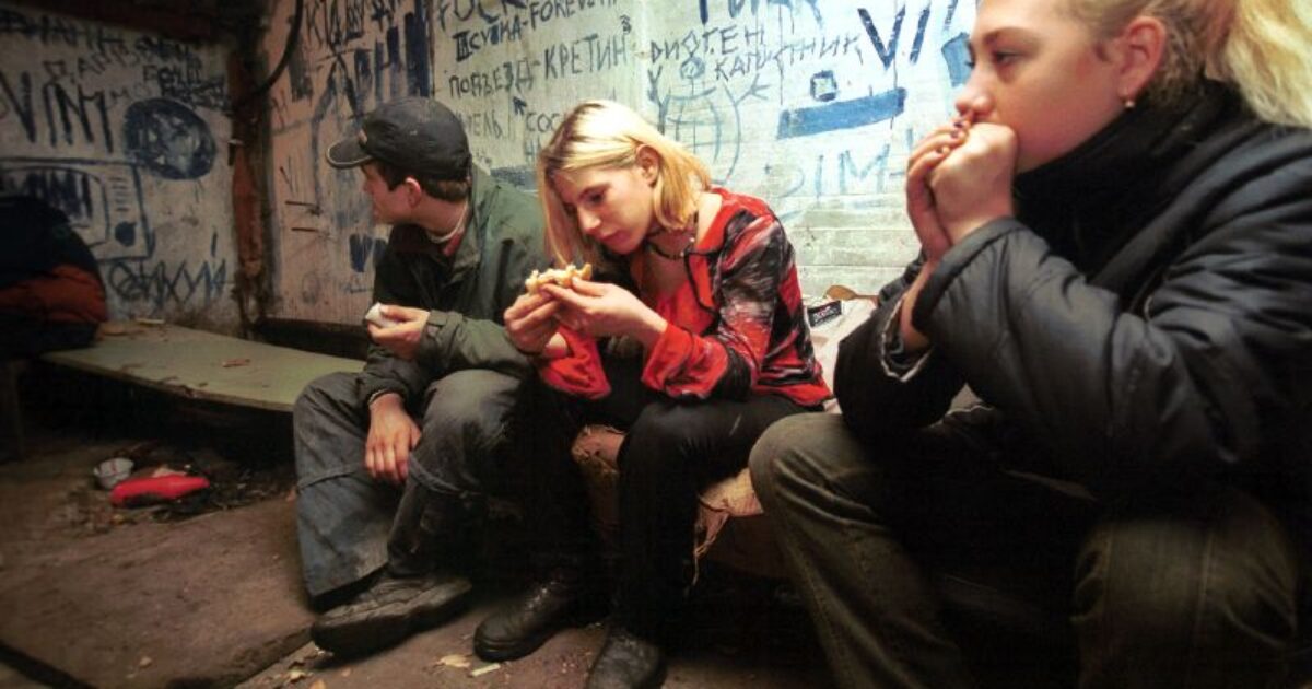 Three young people sit in a dimly lit room with graffiti-covered walls, sharing food and deep in thought.