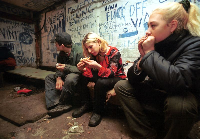 Three young people sit in a dimly lit room with graffiti-covered walls, sharing food and deep in thought.
