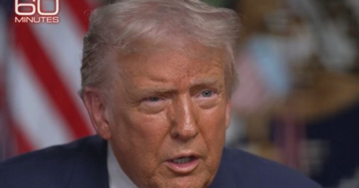 Close-up of a man speaking during a televised interview, with a flag in the background and a serious expression.
