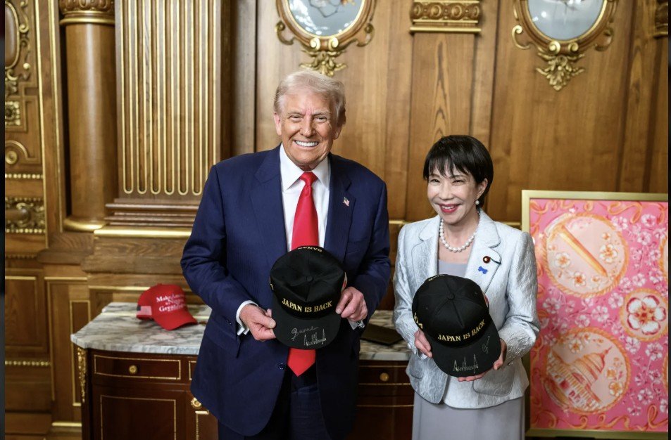 Donald Trump and a woman hold matching "Japan is Back" hats in an ornate room, symbolizing a diplomatic meeting between the U.S. and Japan.