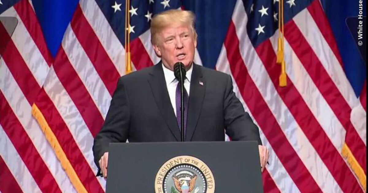 Donald Trump delivering a speech at a podium with the presidential seal, surrounded by American flags.