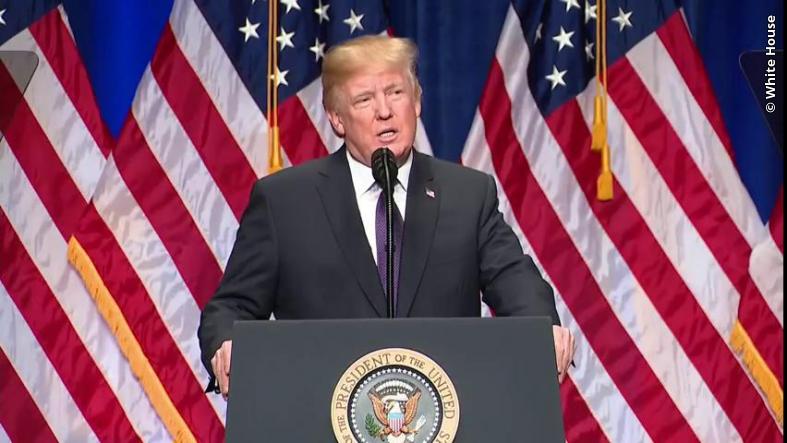 Donald Trump delivering a speech at a podium with the presidential seal, surrounded by American flags.