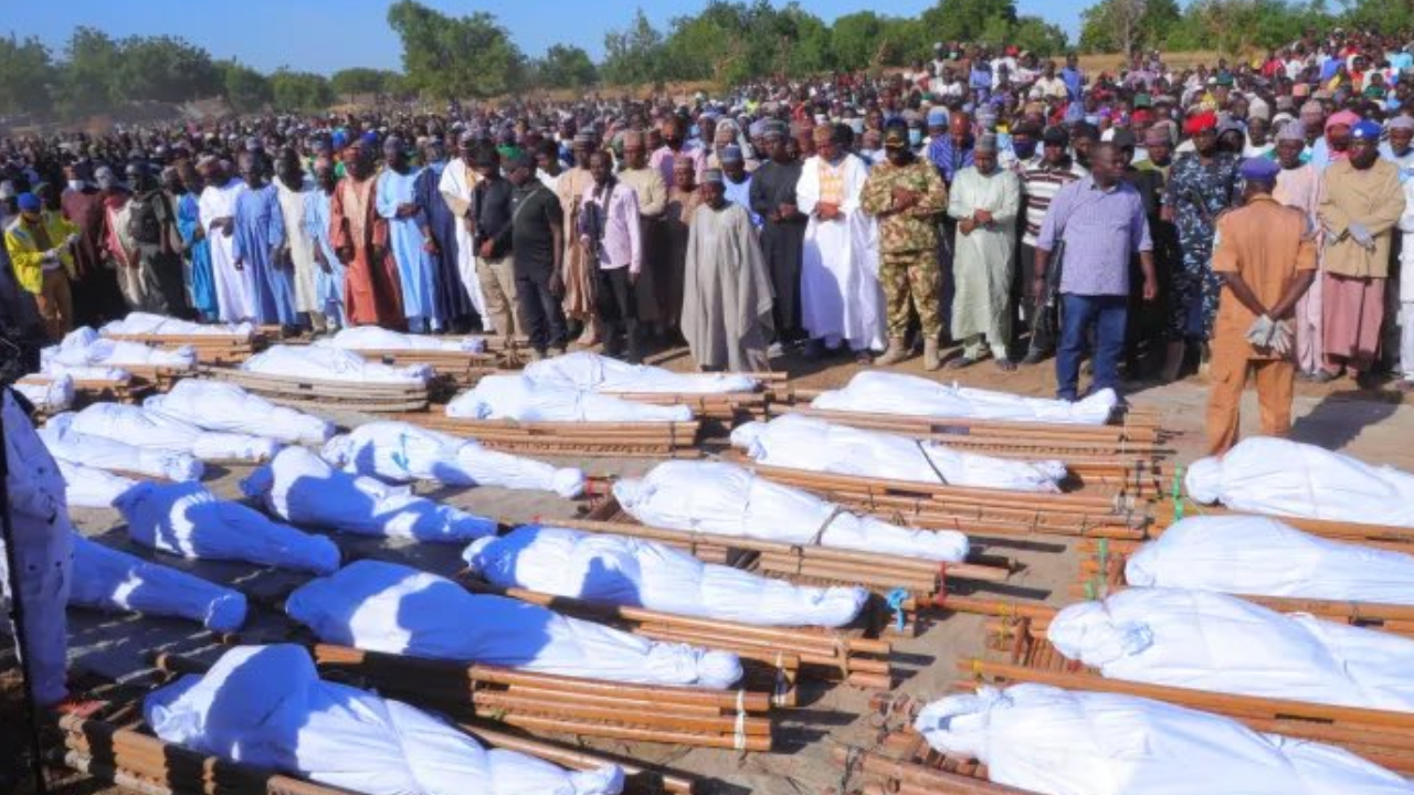 Crowd gathered for a memorial service with white-covered bodies laid out, highlighting a somber moment of mourning and community support.