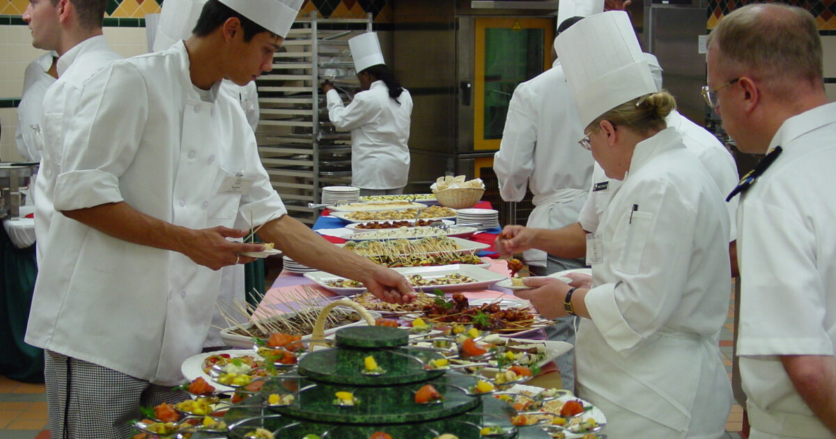Culinary students in white chef uniforms prepare gourmet dishes at a buffet table, showcasing an array of elegantly presented appetizers and entrees in a professional kitchen setting.