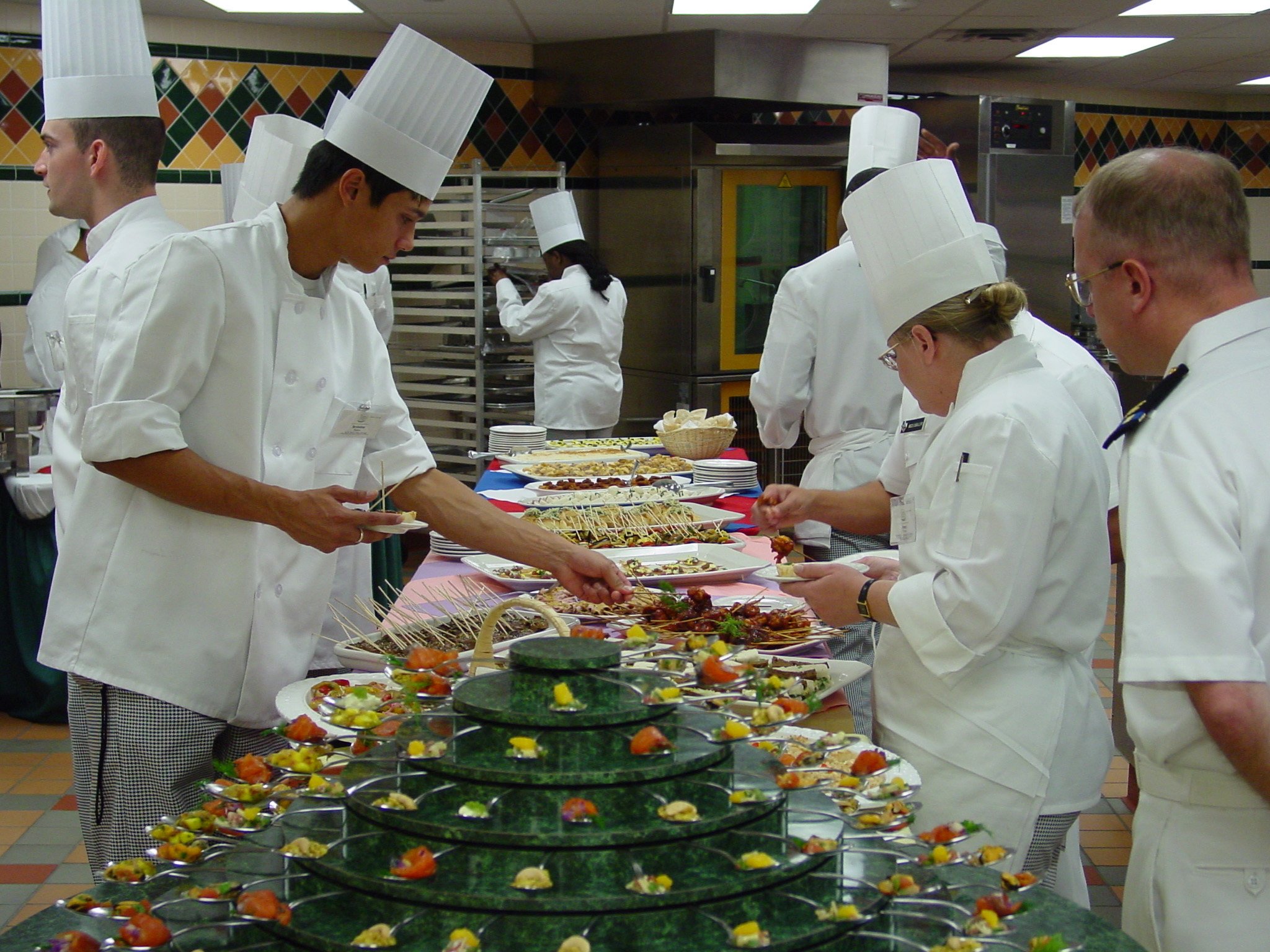 Culinary students in white chef uniforms prepare gourmet dishes at a buffet table, showcasing an array of elegantly presented appetizers and entrees in a professional kitchen setting.
