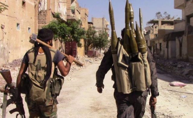 Two armed soldiers walk through a war-torn street, one carrying an axe and the other a backpack loaded with explosive projectiles.