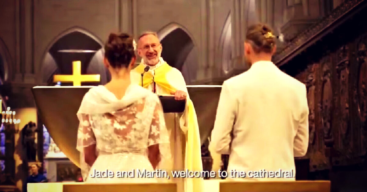 A couple stands before a priest in a cathedral during their wedding ceremony, with a large cross visible in the background.