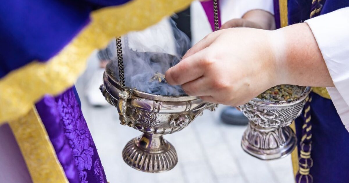 A close-up of a person holding a silver incense burner, releasing aromatic smoke during a ceremonial ritual.