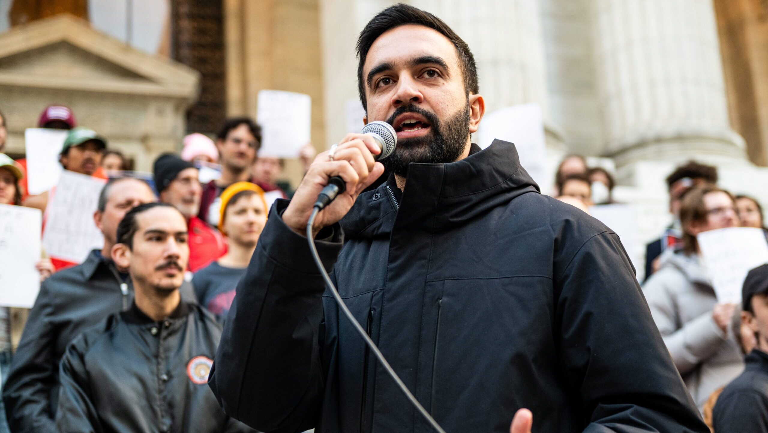 A speaker addresses a crowd during a protest, holding a microphone and advocating for social change outside a historic building.